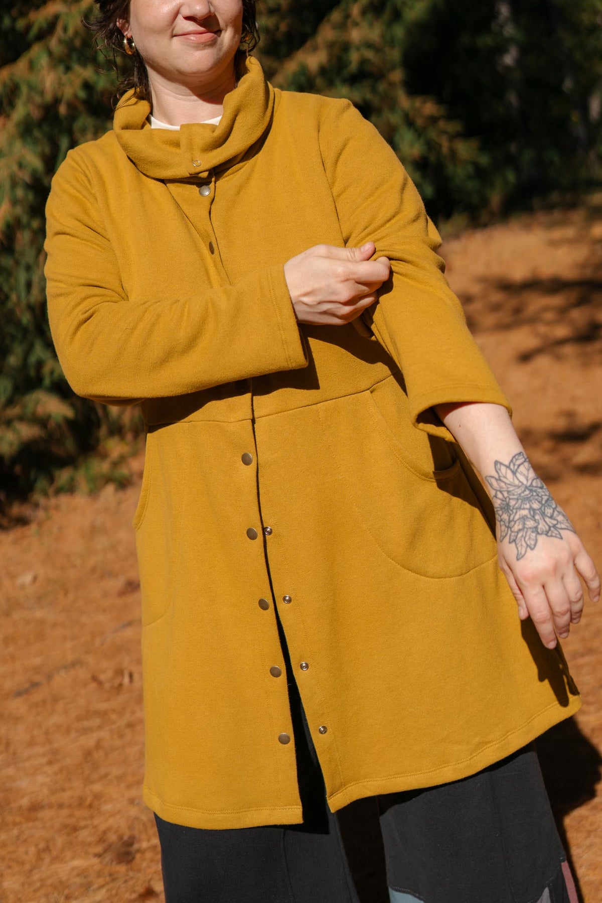 A person wearing a mustard yellow cowl-neck button-front coat outdoors on a dirt path with trees in the background.