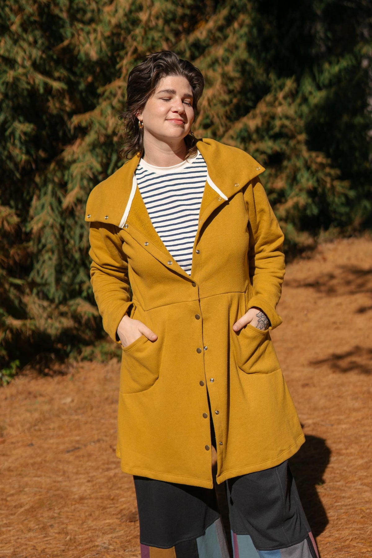 A woman wearing a mustard yellow hooded coat over a black and white striped shirt, standing outdoors in front of a forest background.