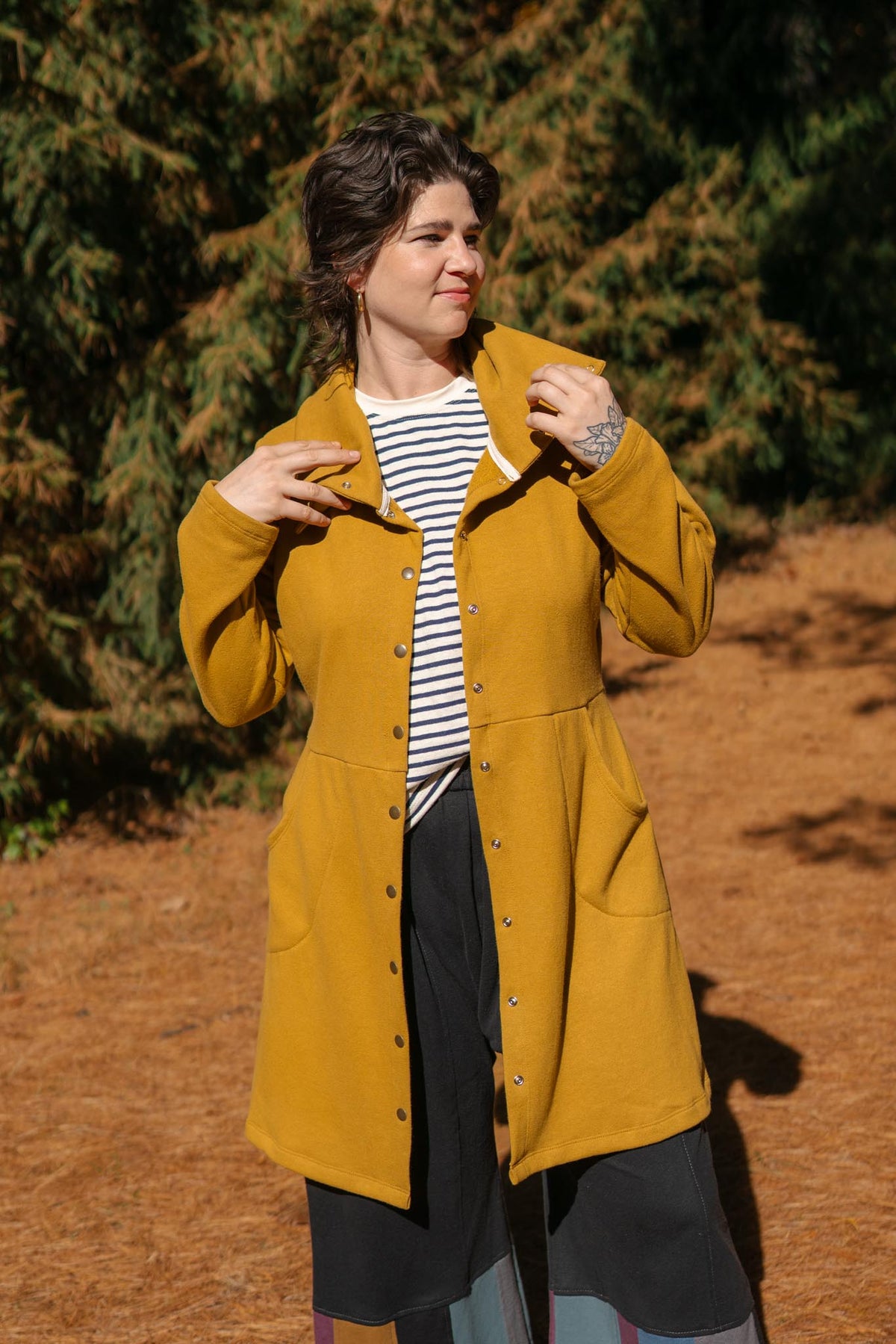 A woman wearing a mustard yellow button-up coat over a black and white striped shirt and dark skirt, standing outdoors on a pine-covered ground with trees in the background.