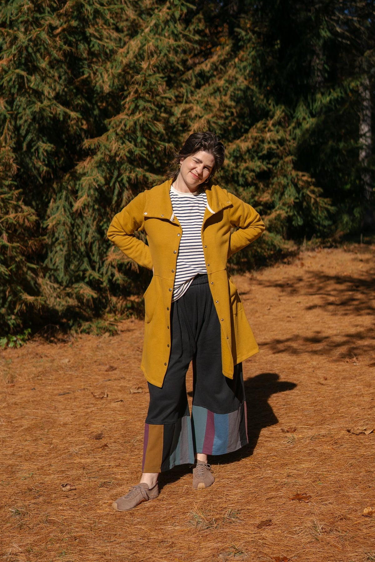 A person standing outdoors on a dirt path in front of evergreen trees, wearing a mustard yellow coat over a black-and-white striped top, dark wide-leg pants, and tan shoes.