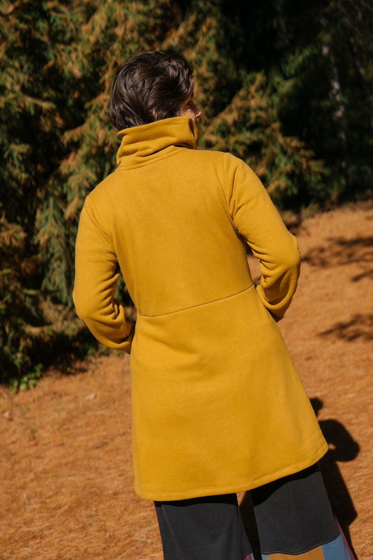 A person viewed from behind wearing a mustard yellow fleece turtleneck coat outdoors on a path with trees in the background.