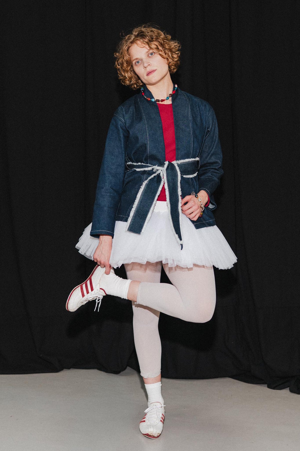 A model poses in a creative outfit featuring a dark blue denim jacket styled like a kimono with a white tie belt, layered over a red top, white tutu skirt, white tights, and white sneakers with red stripes, set against a black curtain background.