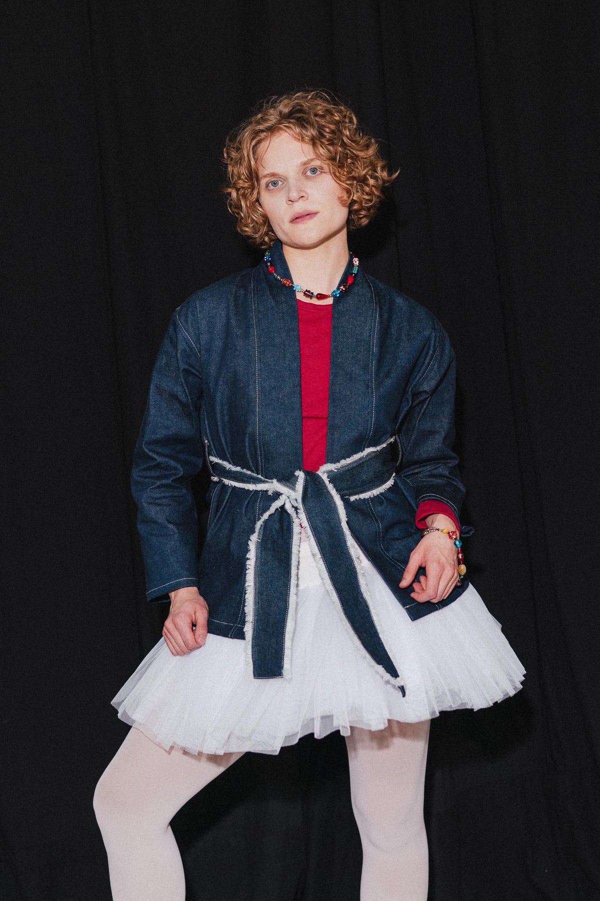 A model with curly light brown hair stands against a black background wearing a dark blue denim jacket with a white frayed tie belt, layered over a red top and a white tulle tutu skirt, paired with white tights.