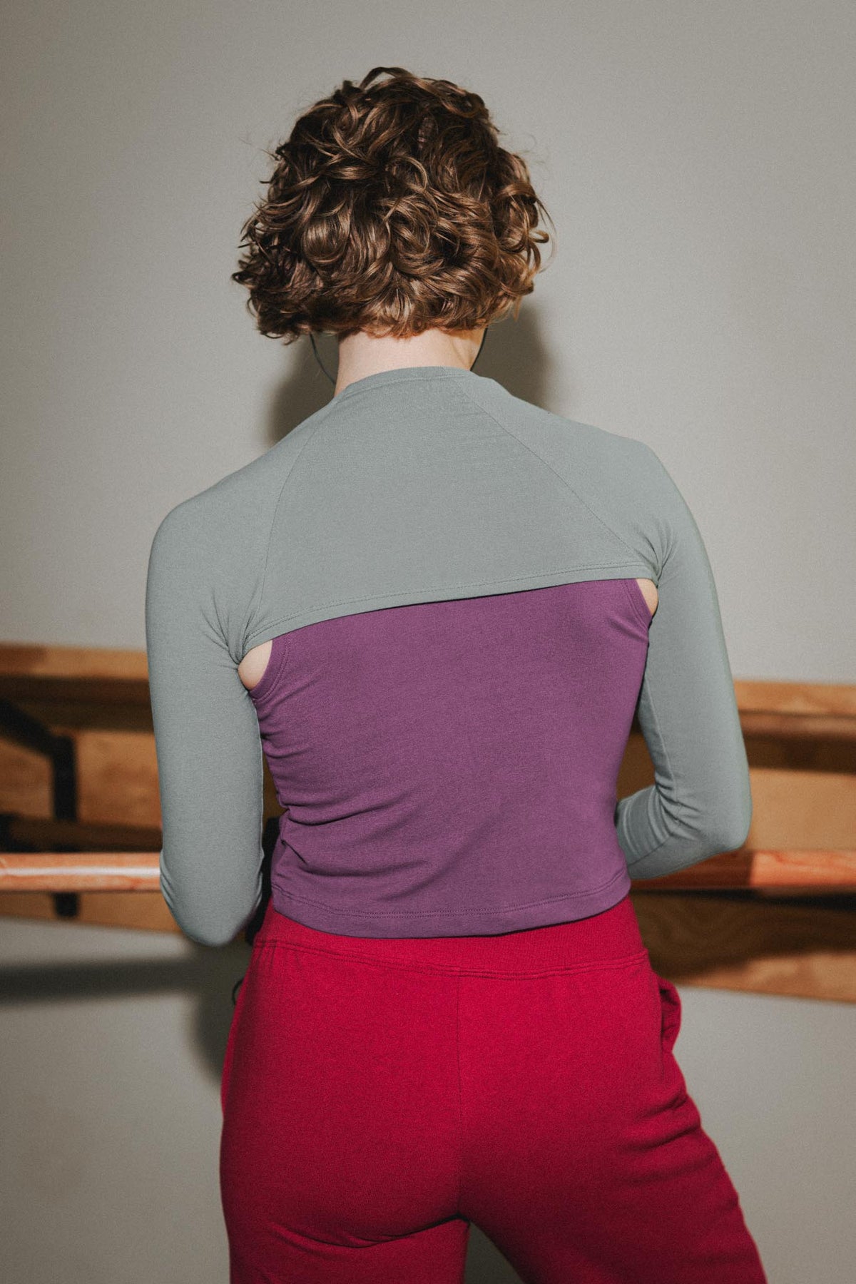 A person shown from behind wearing a color-blocked long-sleeve top with a sage green upper section, a purple middle panel, and red lower pants, standing indoors against a plain wall.