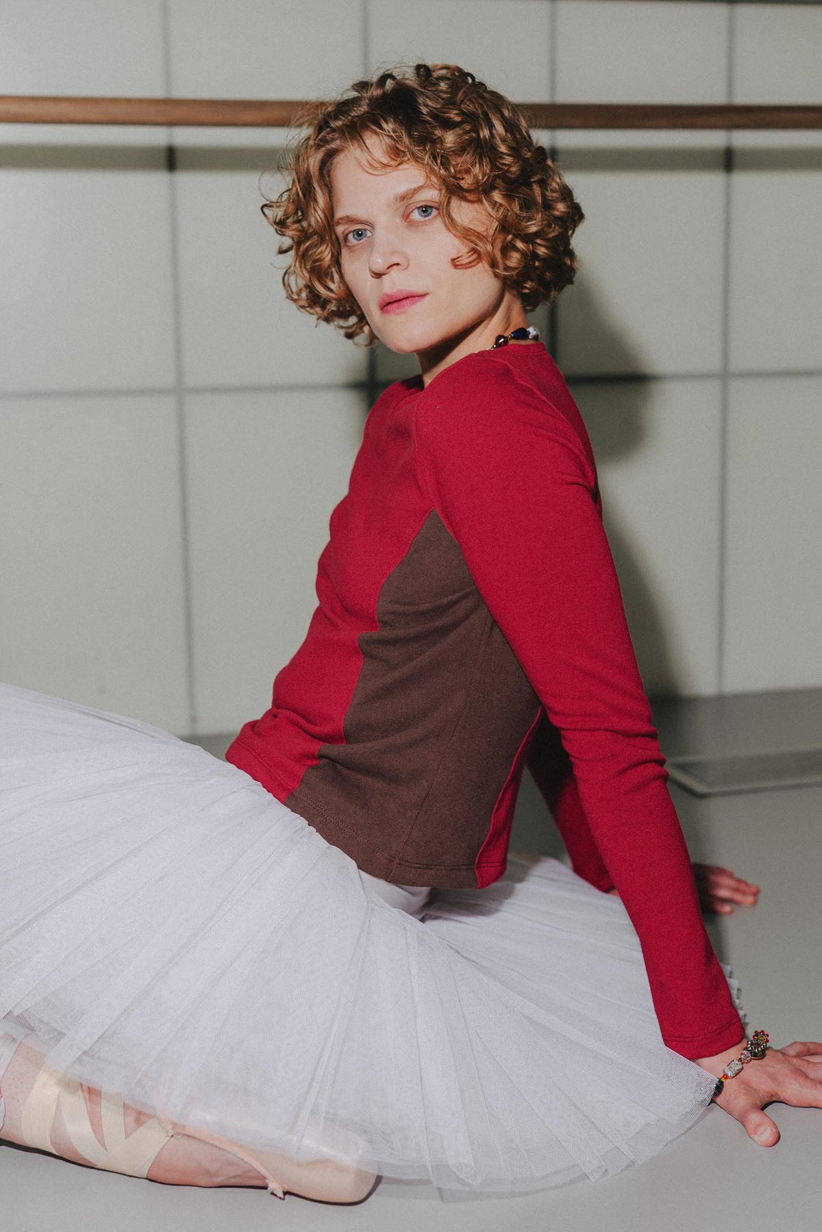 A woman with curly brown hair sits on a ballet barre in a studio, wearing a color-blocked long-sleeve top with red and brown panels, a white tutu skirt, and pointe shoes.