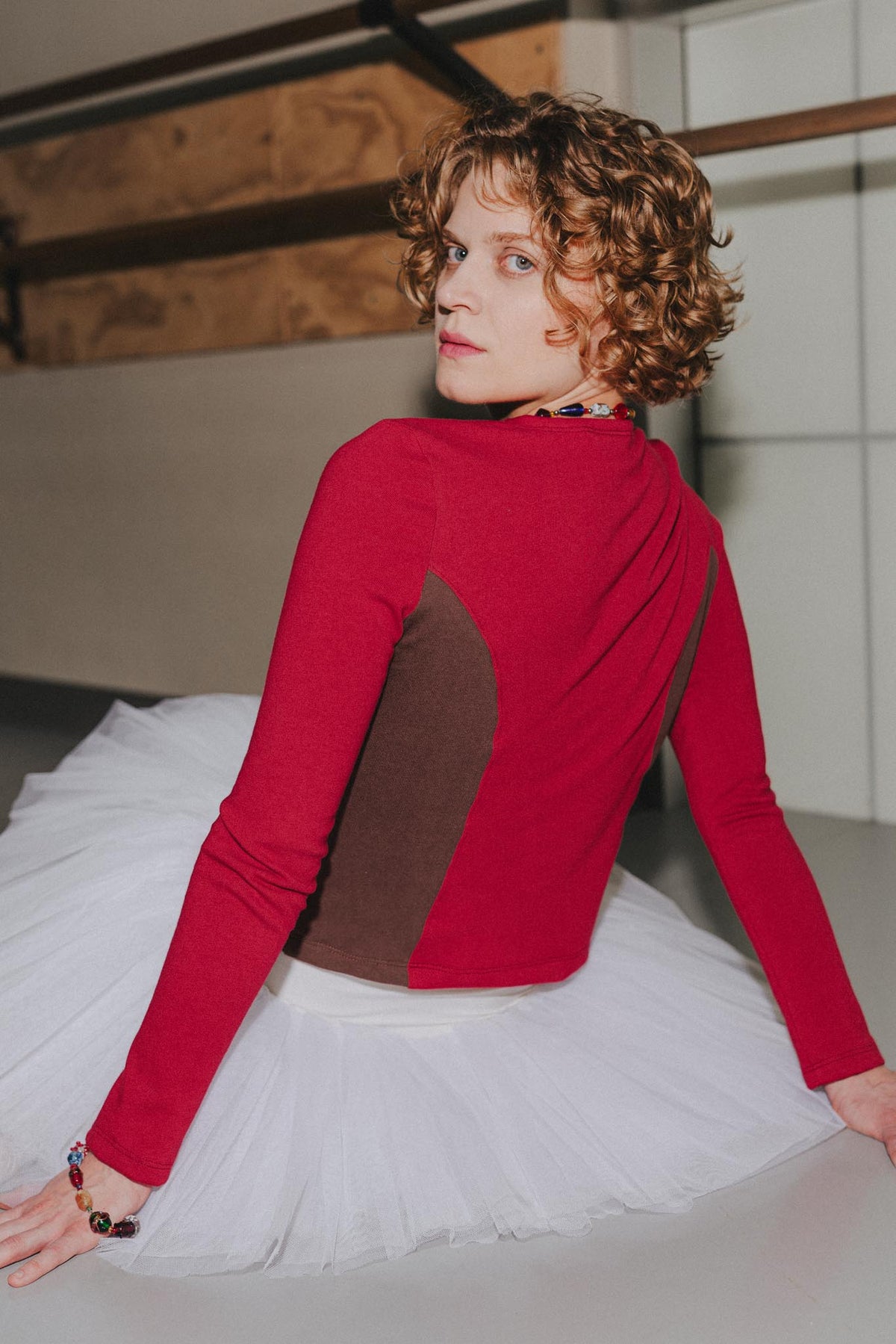 A woman with curly brown hair is shown from behind wearing a red long-sleeve top with a brown color-block panel on the back, paired with a white tutu skirt in a studio setting.