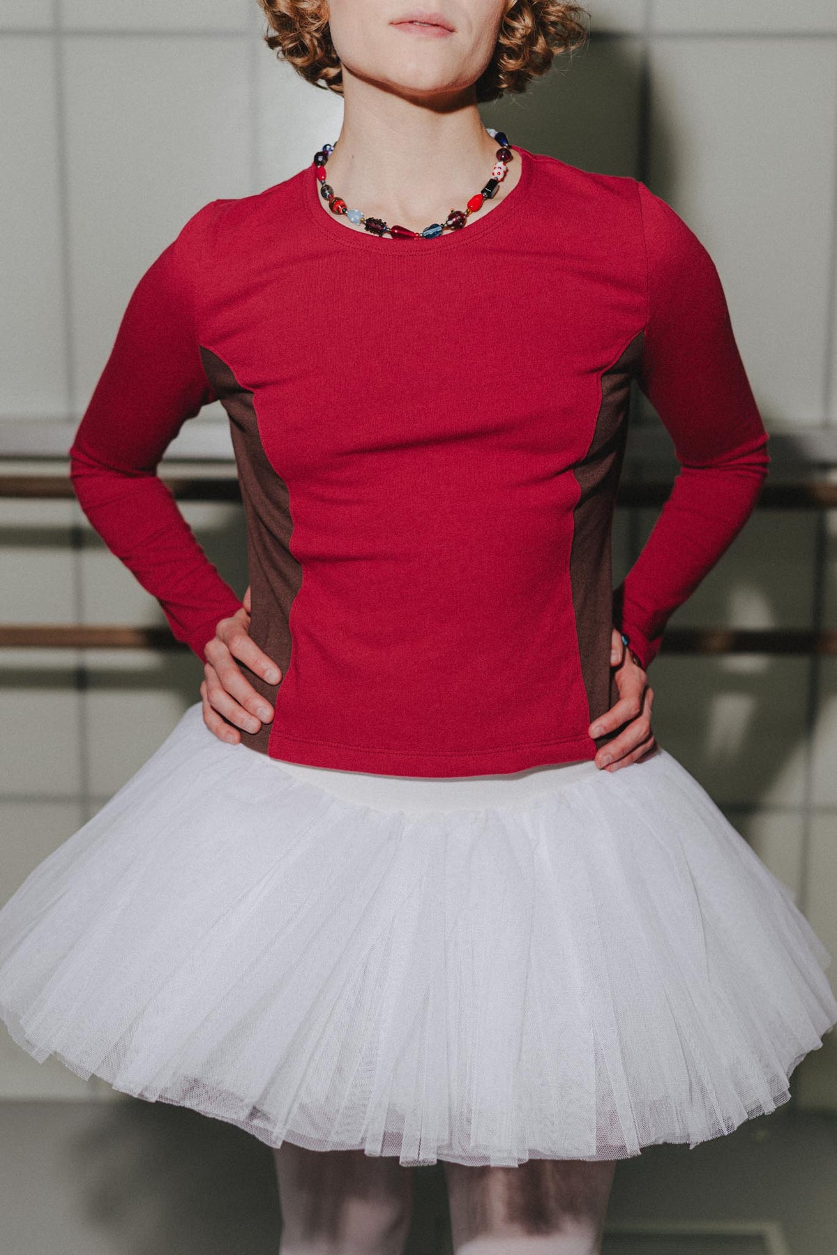A person wearing a fitted long-sleeve top in bright red with dark brown side panels, paired with a white tulle tutu skirt and a chunky beaded necklace.