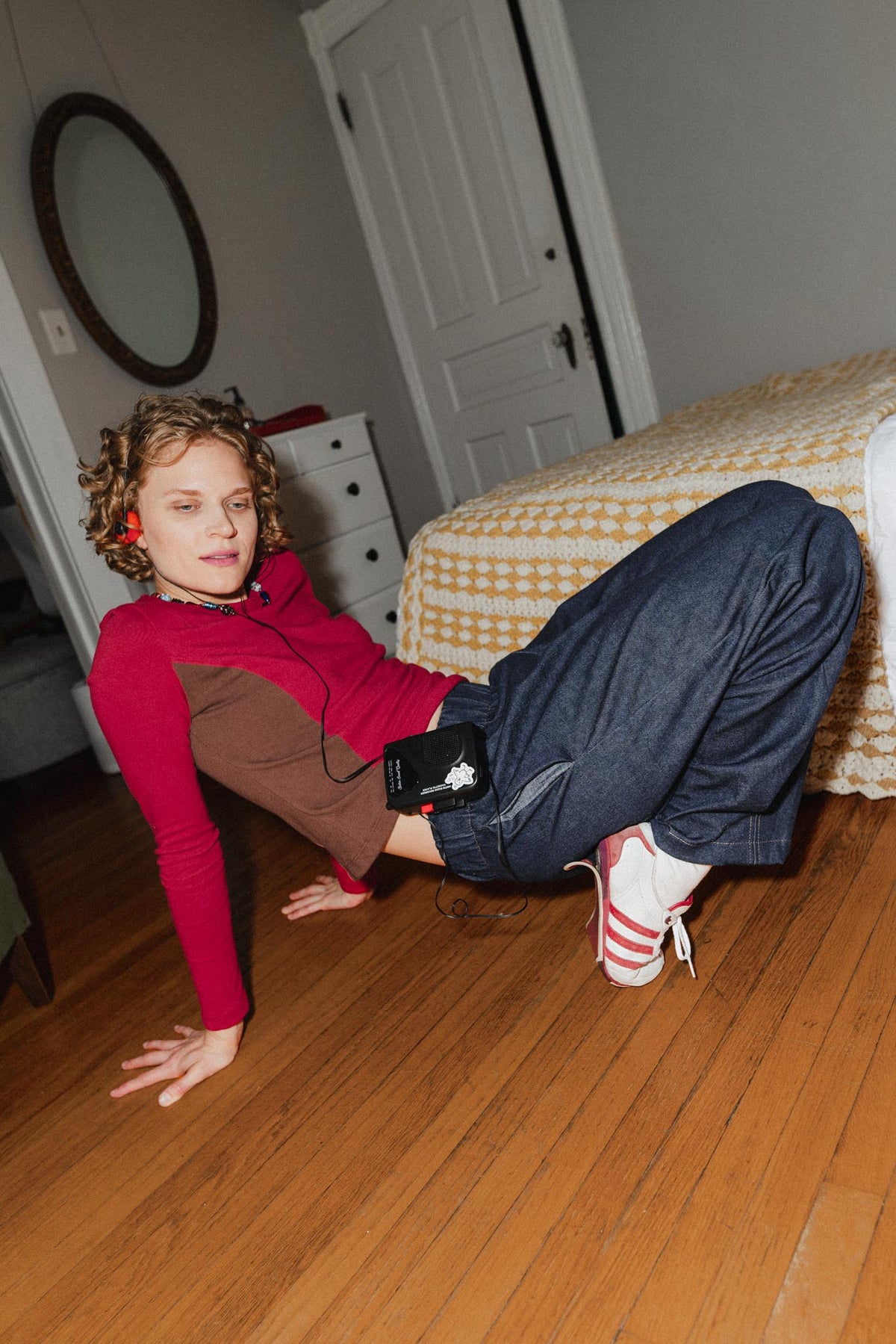 A person in a bedroom setting, wearing a red and brown raglan long-sleeve shirt, dark blue jeans, and white sneakers with red stripes, lying on a wooden floor next to a bed.