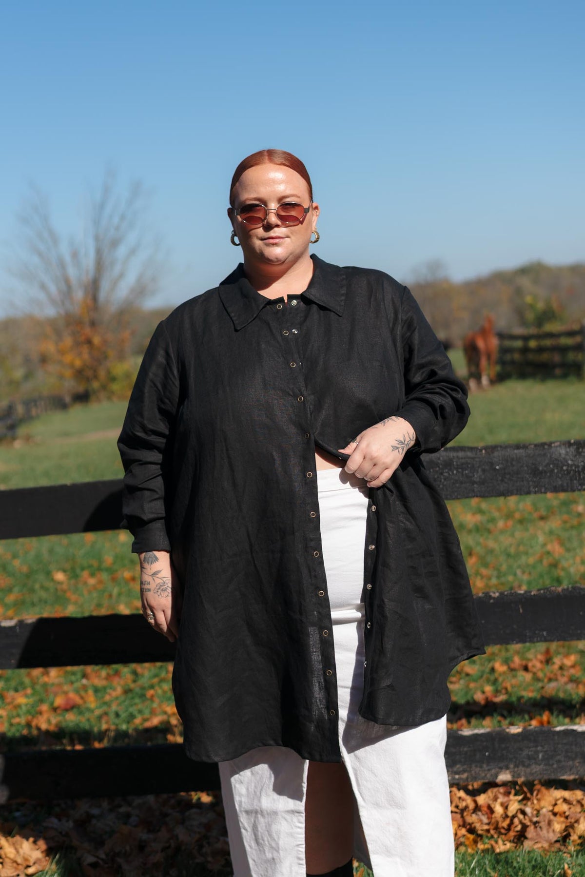 A person standing outdoors in front of a black wooden fence, wearing a long black button-front overshirt over a white top and white pants, with reddish hair and sunglasses.