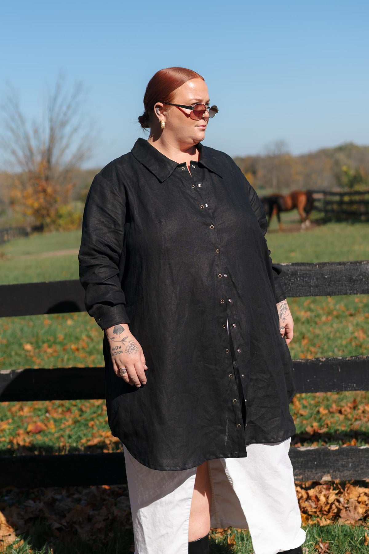 A person wearing a loose black long-sleeve button-front shirt dress or tunic with white pants, standing outdoors in front of a black wooden fence with a grassy field and trees in the background.