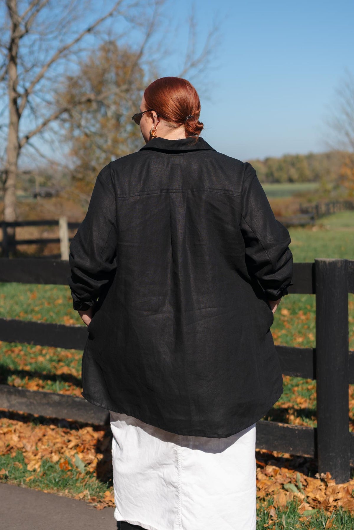 A person viewed from behind wearing a loose black linen shirt and a white skirt, standing outdoors in front of a wooden fence with fallen leaves and trees in the background.