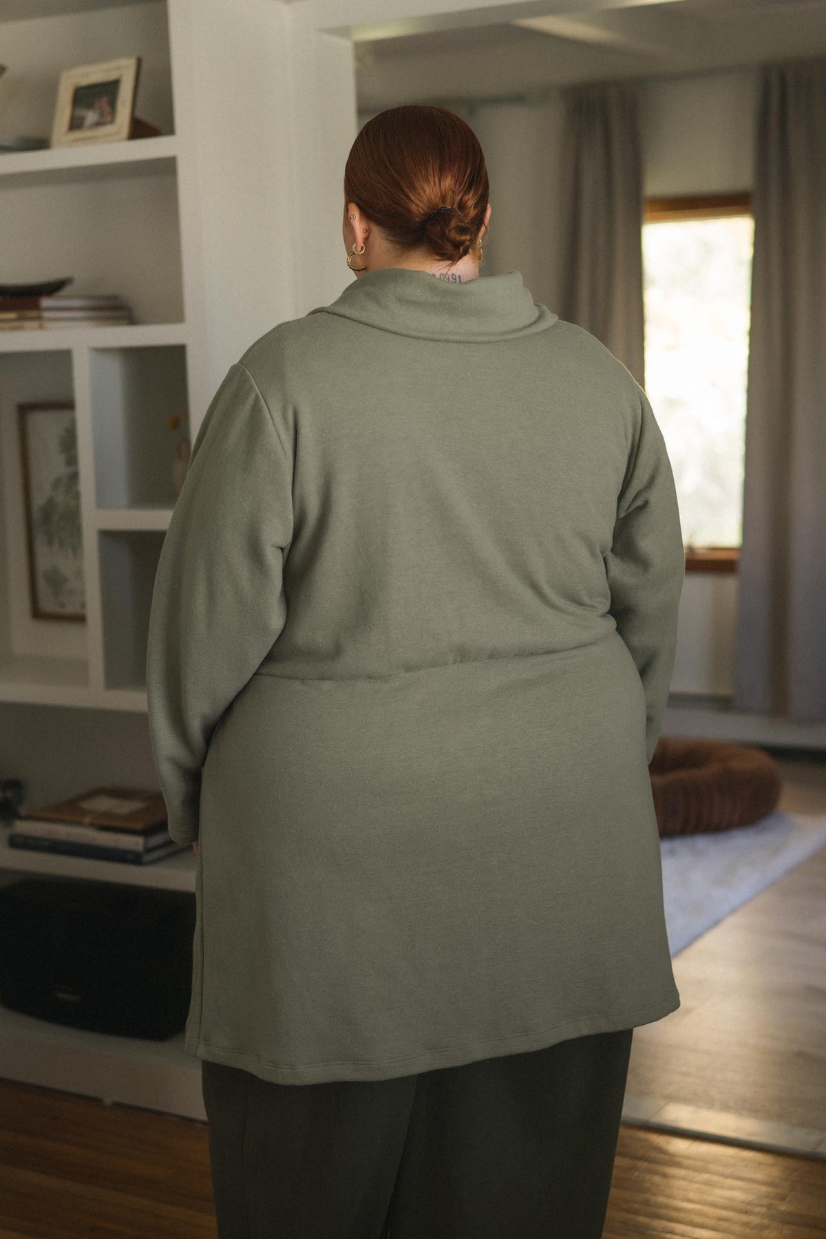 A person viewed from behind wearing a muted sage green long-sleeve top and dark pants in a softly lit indoor room with shelves, curtains, and a window.