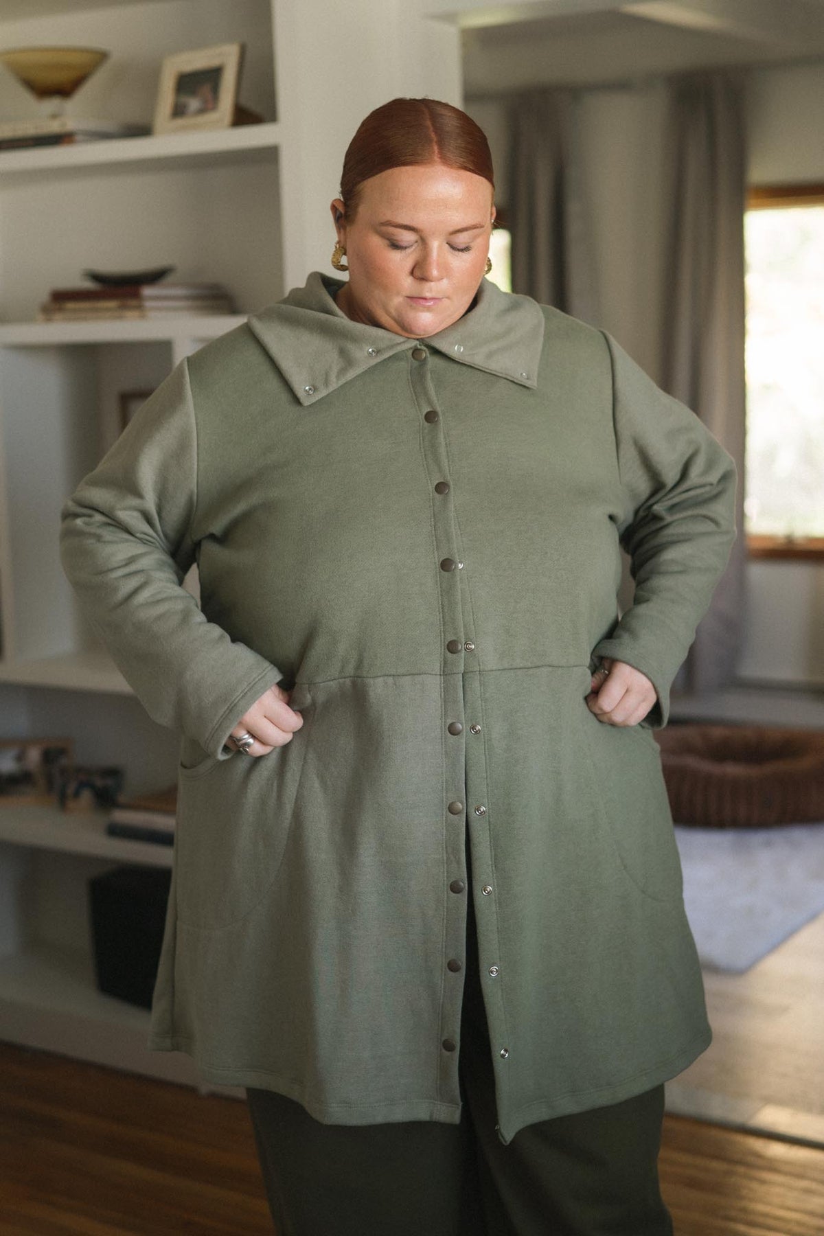 A woman wearing a muted sage green button-front tunic or coat with a wide collar and front pockets, standing in a home interior with shelves and curtains visible in the background.