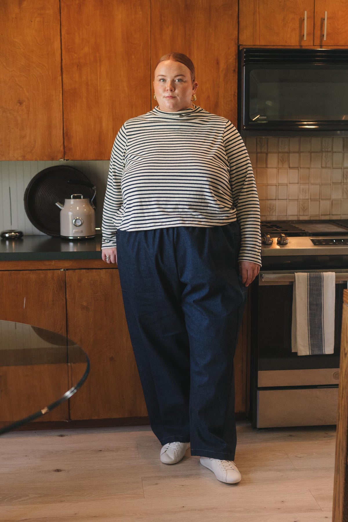 A woman stands in a kitchen wearing a striped long-sleeve mock-neck top in navy and white, paired with loose dark blue pants and white sneakers.