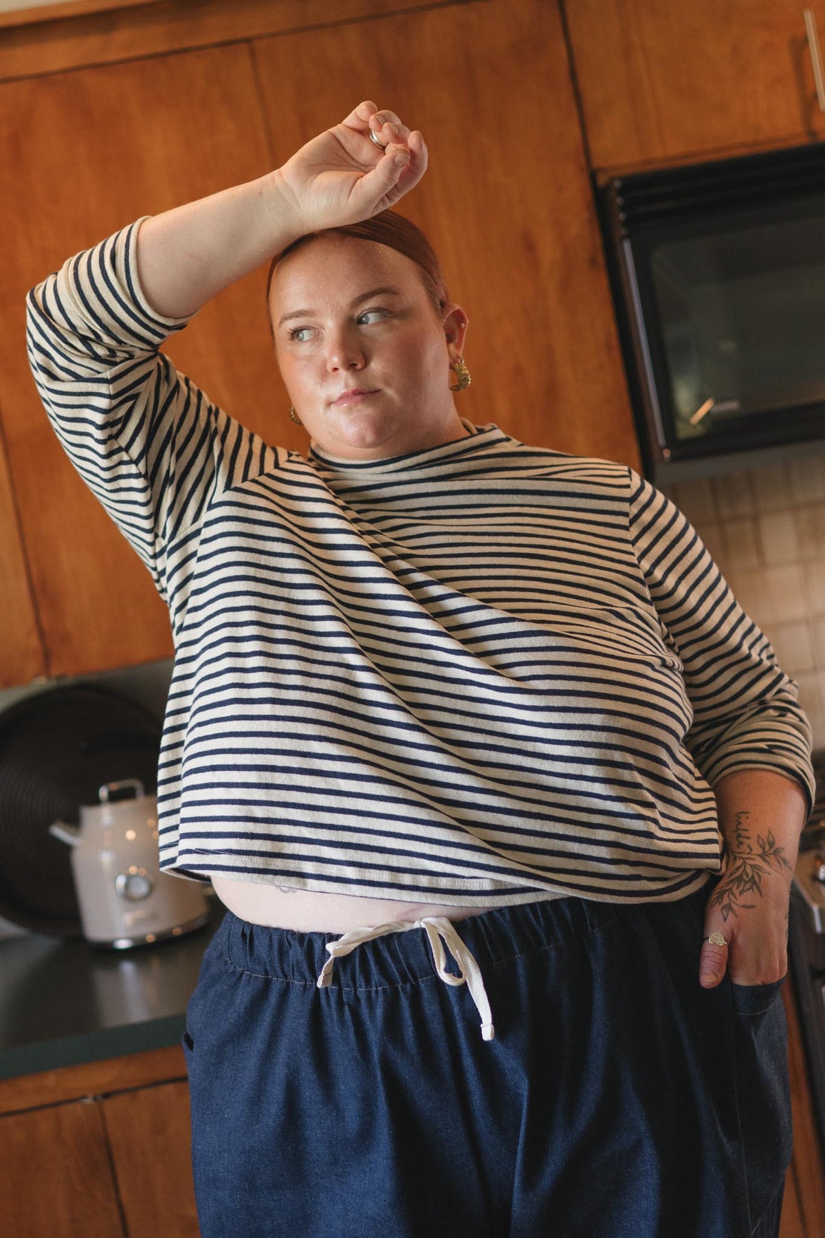 A person standing in a kitchen wearing a navy and white horizontally striped long-sleeve top and dark blue drawstring pants, with one arm raised to their head.
