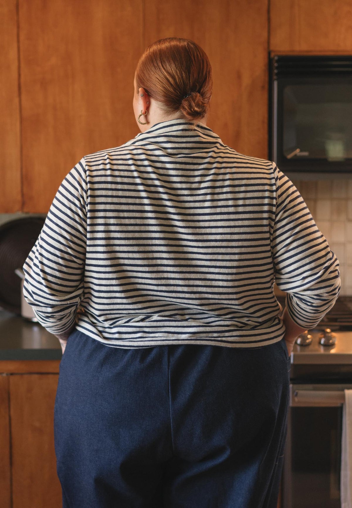 A person viewed from behind wearing a navy and white horizontally striped long-sleeve shirt and dark blue pants in a kitchen setting with wooden cabinets.