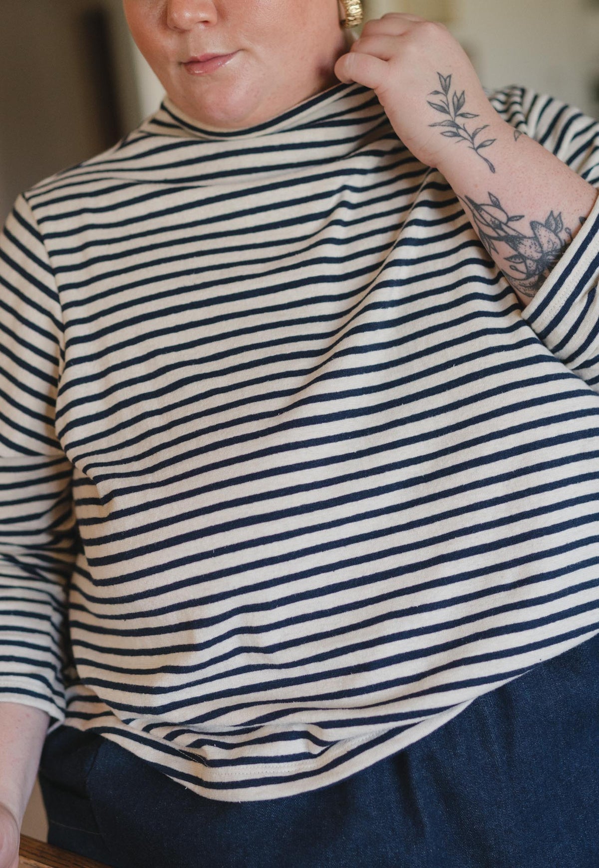 A close-up of a person wearing a navy and cream horizontal striped turtleneck top, with a floral tattoo visible on their left arm.