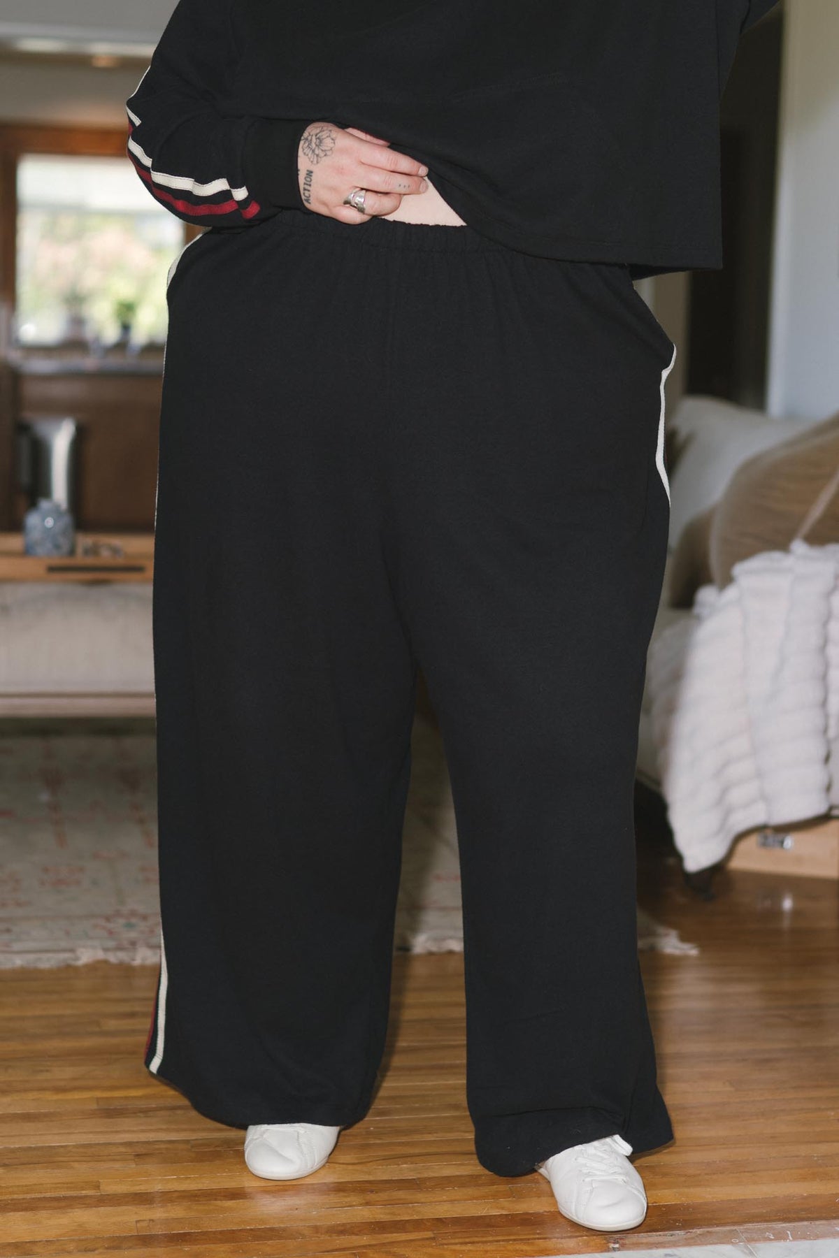 A person wearing black wide-leg sweatpants with white side stripes, paired with a black sweatshirt and white sneakers, standing in a living room.