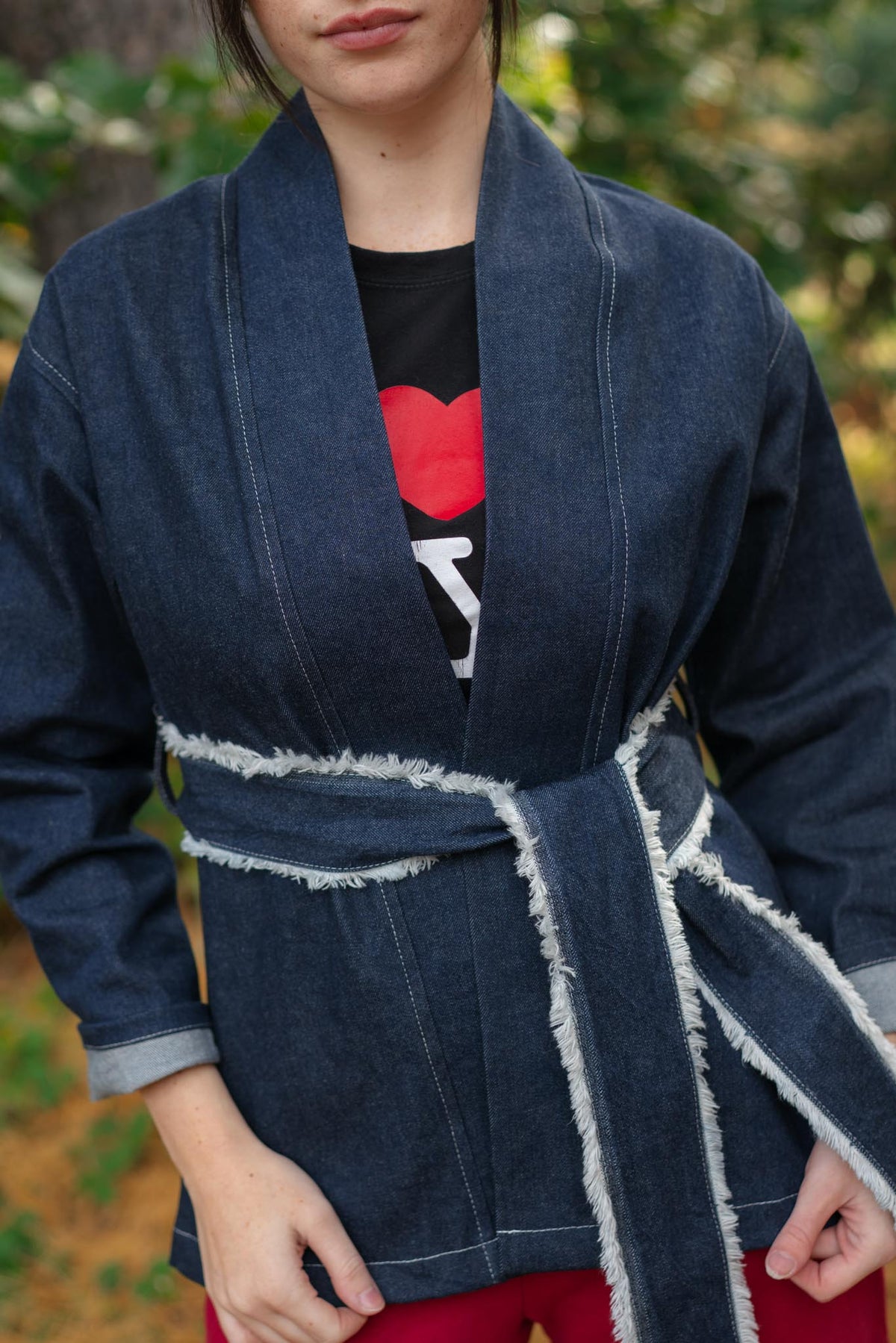 A person wearing a dark indigo denim kimono-style jacket with raw, frayed edges and a matching tie belt, layered over a black graphic t-shirt and red pants, set against a blurred outdoor background.