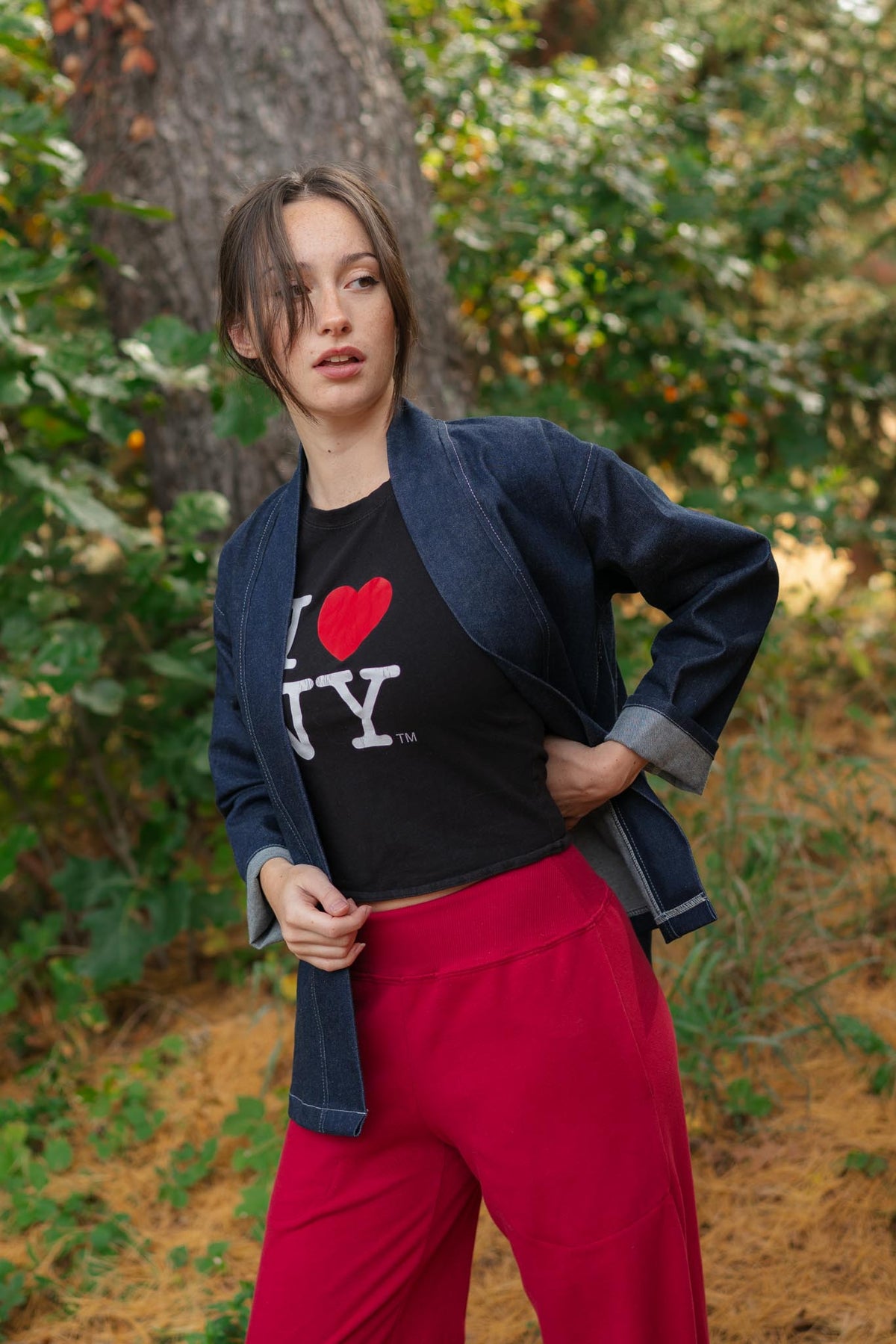 A woman posing outdoors in a wooded setting, wearing a black graphic t-shirt with a red heart and white 'NY' text, a dark blue open-front jacket, and bright red pants.