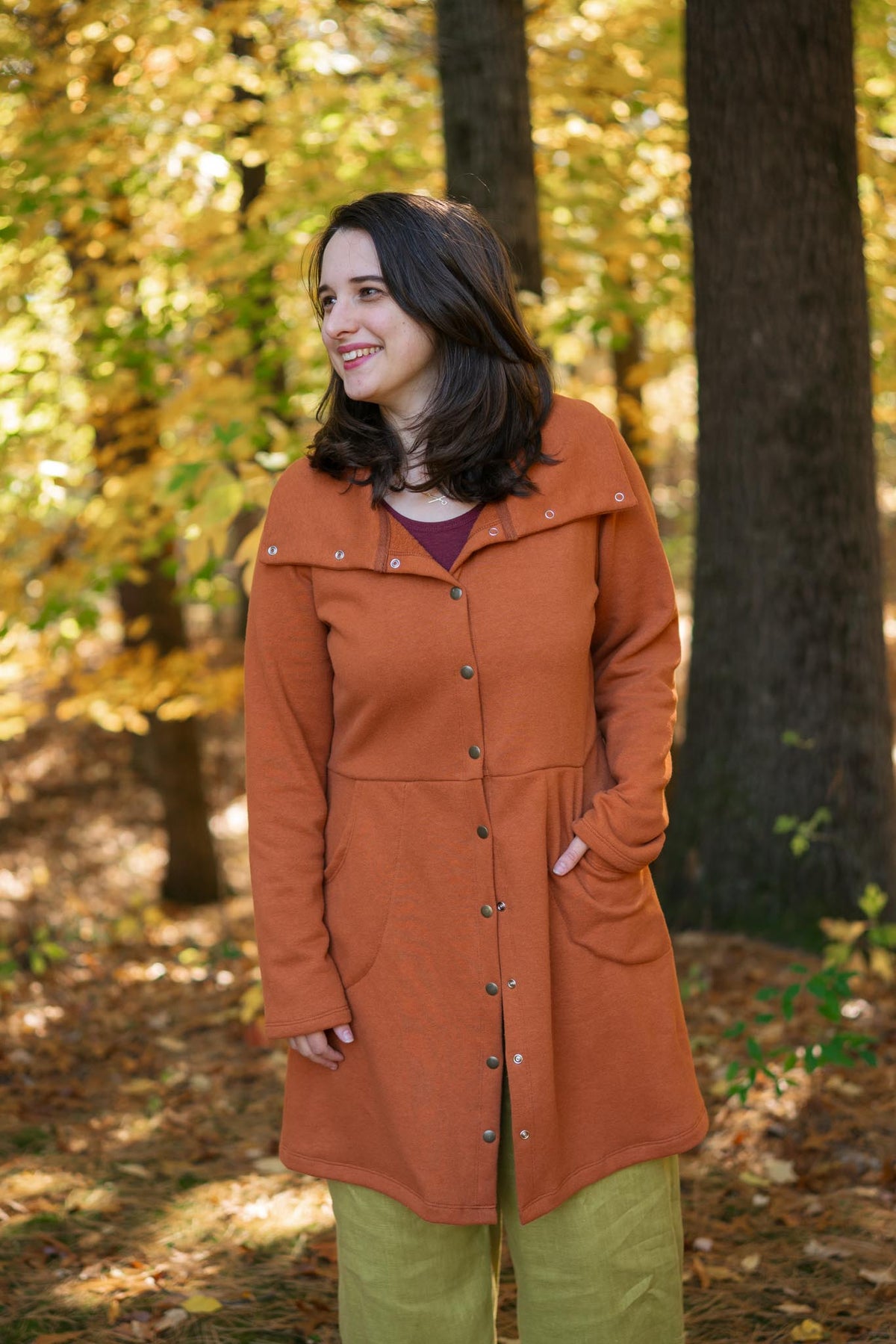 A woman wearing a rust-orange mid-length button-front coat with a wide collar and olive green pants, standing in a sunlit autumn forest with trees and fallen leaves.