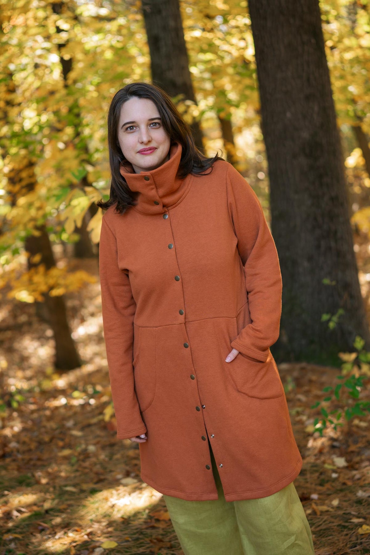 A woman wearing a rust-orange mid-length button-front coat with a high mock neck and olive green pants, standing in an autumn forest with trees and fallen leaves in the background.