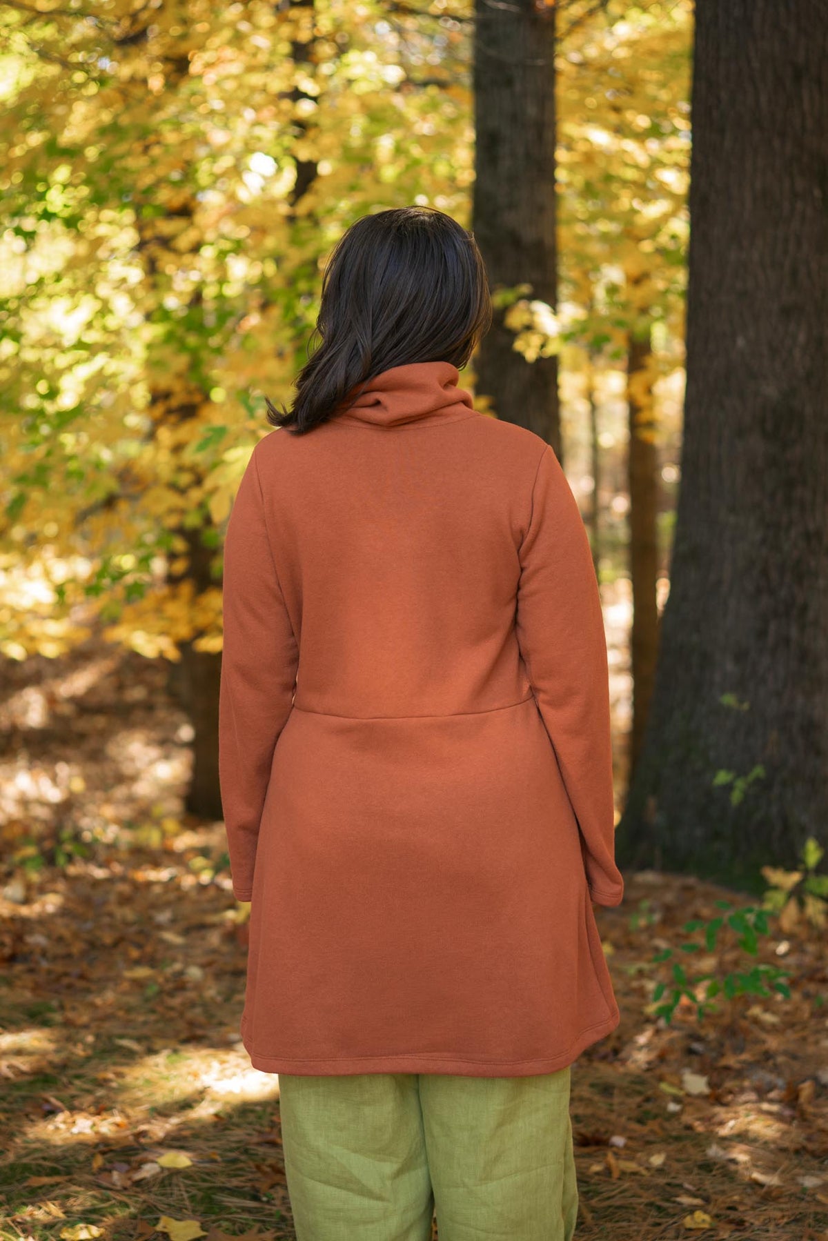 A woman viewed from behind wearing a rust-orange long-sleeve tunic coat and light olive green pants, standing in a sunlit autumn forest with yellow leaves and tree trunks.