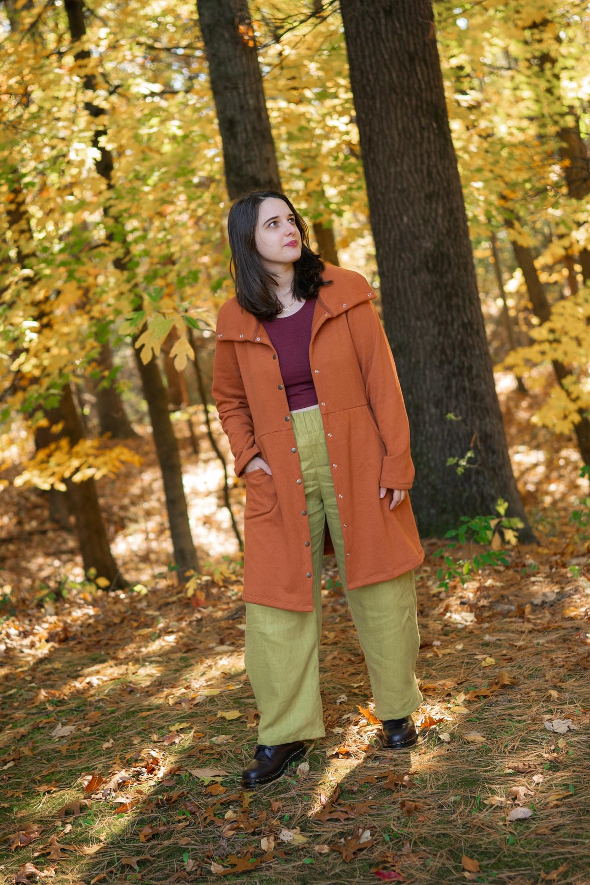 A woman standing in a wooded autumn setting, wearing a rust-orange mid-length coat over a dark plum top and light olive green wide-leg pants.
